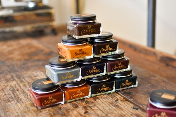 A vibrant pyramid of Saphir shoe polish jars on a rustic wooden table, showcasing the rich colors and quality.