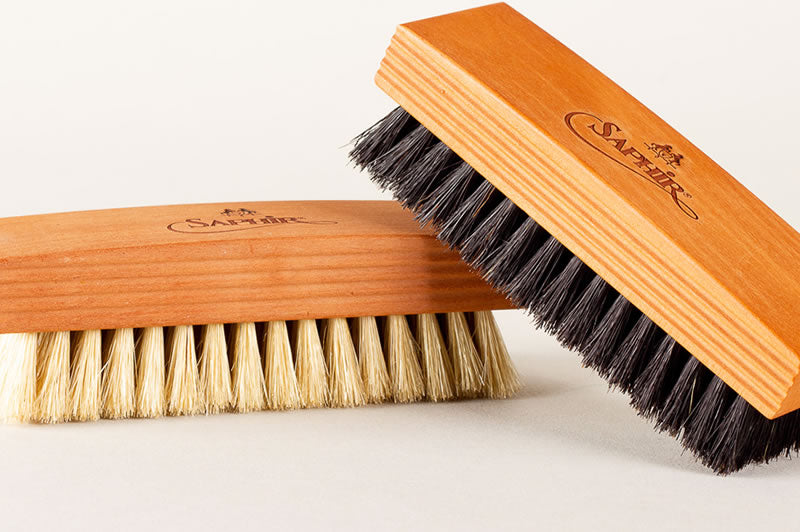 Two wooden-handled brushes with bristles: one has dark bristles and rests at an angle atop another with light bristles. Both handles are engraved with the Saphir logo.